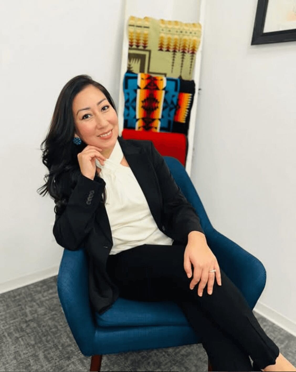 A professional portrait of Monique Castro, a Diné (Navajo) Nation and Xicana therapist and leadership coach, sitting confidently in a blue chair. She is wearing a black blazer and white blouse, with a warm smile. A colorful Indigenous blanket hangs in the background.