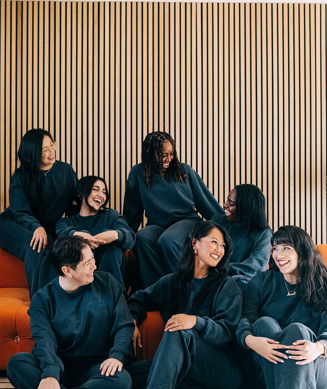A diverse group of seven women sitting and lounging on an orange couch, all dressed in matching dark-colored sweatshirts and sweatpants. They are laughing and engaging with each other, with a warm and friendly atmosphere. The background features a wooden slat wall.