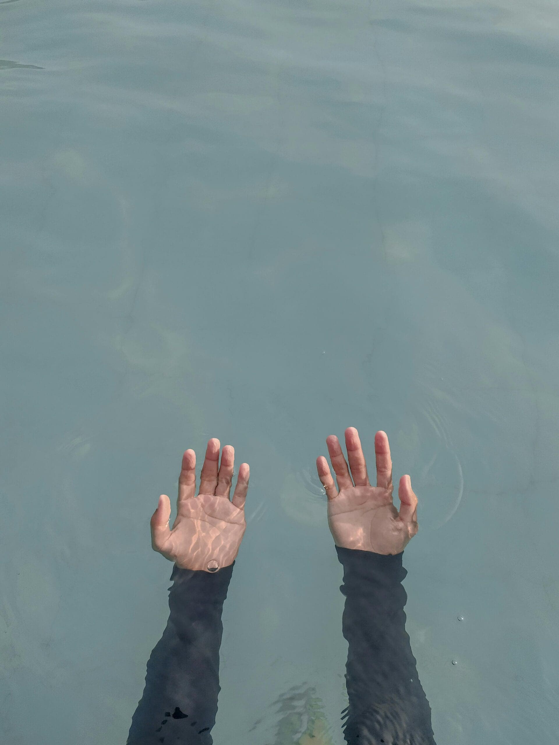 Hands underwater reaching above the surface, the individual is wearing sleeves as if they are swimming with all their clothes on